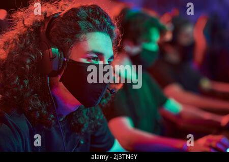 Curly-haired young Caucasian man in cloth mask and headphones participating in esports competition Stock Photo