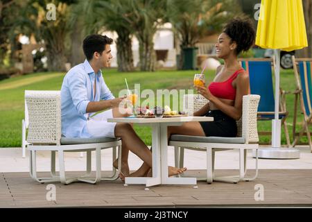 interracial couple of young people eating a fruit breakfast relaxing sitting on the backyard table. Multiracial young lovers at the resort talking and Stock Photo