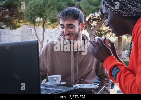 biracial couple of young people using laptop sitting at outdoors cafe and drinking cappuccino Stock Photo
