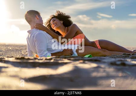A young multiracial romantic couple tenderly caresses each other lying on the beach at sunset - romance multiracial couple on the beach - Love and tra Stock Photo