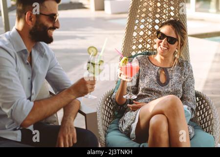 Couple of friends with sunglasses talking and flirting in the terrace garden holding cocktails - Young people toasting together Stock Photo