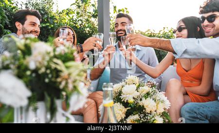 Friend sparty gathering in the terrace and toasting champagne sitting in the couch - best friends having fun drinking sparkling wine in the luxury gar Stock Photo