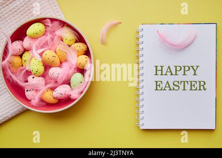Happy Easter text. Pink bowl with colorful eggs and pink feathers. Happy Easter concept. Post card on bright yellow background Stock Photo