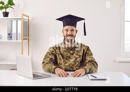 Happy young student in camouflage uniform and graduate hat sitting at desk and smiling Stock Photo