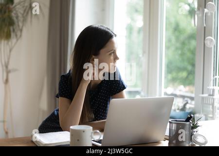 Thoughtful woman sit at table with laptop looks out window Stock Photo