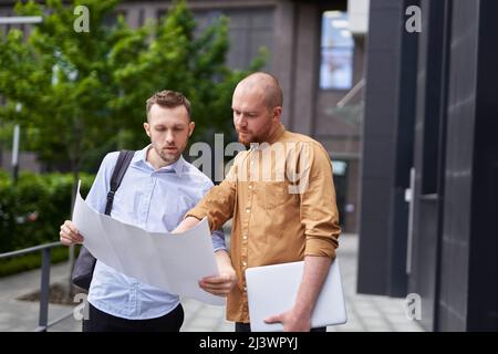 Business team talking outdoor near modern building background. Male caucasian architects or designers holding drawings or blueprints discussing project, brainstorming. High quality image Stock Photo