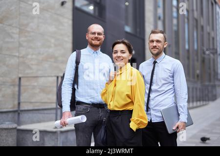 Confident young confident business colleagues standing smiling outside office building looking forward with blueprints and laptop. Team of economists or real estate managers in casual clothes outdoors Stock Photo