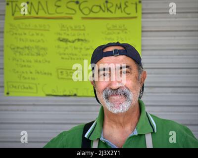 Positive Mexican man (cook, chef) sells traditional food (tamale) at the slow food market, stands in front of hand written menu and looks at viewer. Stock Photo
