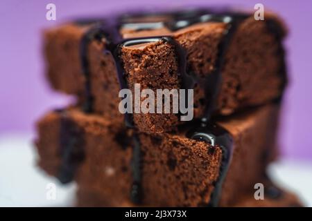 Very close shot of a Pile of Chocolate Brownies with chocolate strands falling on a white plate with purple background. Stock Photo