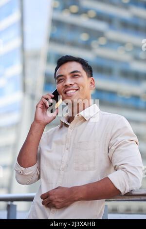 An Asian businessman stands on a city office balcony talking on his cellphone Stock Photo