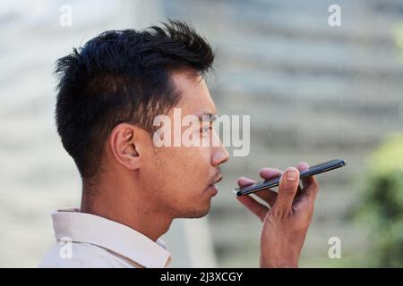 A young Asian man sends a voicenote using cellphone with city in background Stock Photo