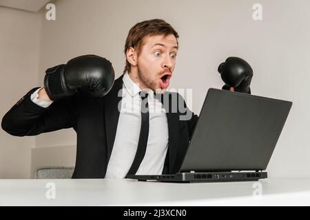 Young angry businessman in black suit wearing boxing gloves using laptop. Stock Photo