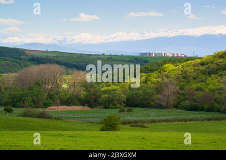 Wonderful springtime landscape, grassy field and rolling hills. Snow-capped high mountains. Buildings from a rural town in the distance. Stock Photo