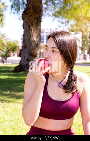 Close-up of a young Caucasian woman dressed in a top and leggings is sitting on the grass of a park while eating an apple. Stock Photo