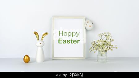 Frame and flowers on background of white table and wall. Eggs and fun rabbit with golden ears. Happy Easter. Decor Stock Photo