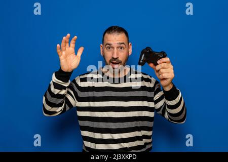 Photo of funny attractive surprised man holding hands with joystick playing video game, excited gamer riding speed car to finish wearing striped Stock Photo