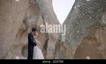 Beautiful young wedding couple in Cappadocia mountains under romantic snowfall. Action. Bride and groom together standing in the gorge surrounded by Stock Photo