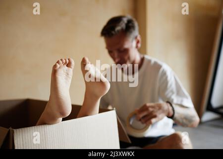 Close up of cute baby feet sticking out of cardboard box while family moving house in background, copy space Stock Photo