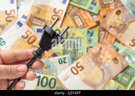Woman hand holding black power plug over euro banknotes background. Power crisis and inflation. Concepts of increasing the energy costs, heating costs Stock Photo