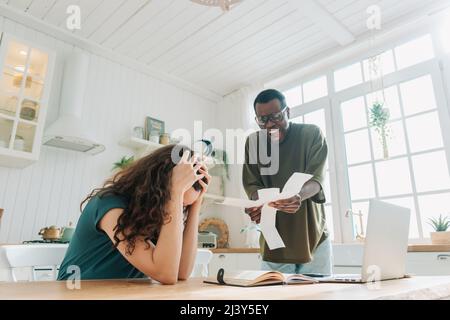 African American man scolds white woman for spending too much money on shopping showing checks. Multinational couple quarrels in kitchen at home Stock Photo