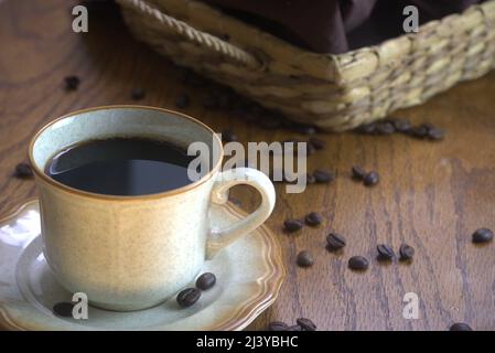A cup of black coffee on a brown rimmed saucer with 3 coffee beans on it. Spilled coffee beans are on the brown textured tabletop next to a wicker tray. Stock Photo