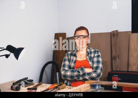 Portrait of a young red-haired female carpenter in her small carpentry workshop. Looking at the camera Stock Photo