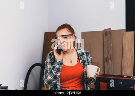 Young red-haired female carpenter talking to a customer on her mobile phone, in her small workshop. Stock Photo