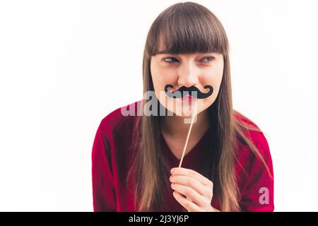 Pretty young woman holds fake moustache in front of her face and looks away movember concept white background copy space isolated studio shot . High quality photo Stock Photo