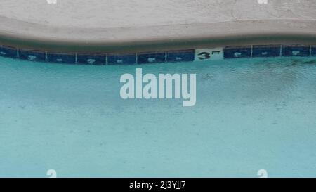 Rain drops falling on water surface of blue swimming pool, rainy day in motel or hotel, California USA. Rainfall on summer vacations, raindrops splashing during monsoon. Seamless looped cinemagraph. Stock Photo