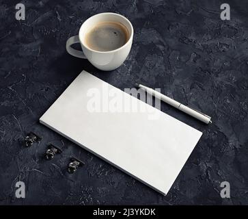 Blank white paper envelope, coffee cup, pen and metal clips on black plaster background. Stock Photo