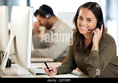 Take note of how expert customer care should be done. Portrait of a young businesswoman using a headset and writing in a notebook in a modern office. Stock Photo
