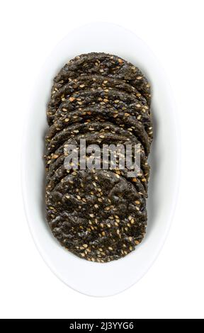 Sesame crackers, in a white bowl. Japanese senbei, crispy macrobiotic crackers, made of brown rice, black sesame seeds and soy sauce. Crunchy snack. Stock Photo