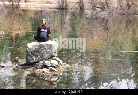 Yosemite National Park, CA - 26 March 22: Young female hiker sits on a rock at Mirror Lake. Trees and rocks are reflected in the still water. Stock Photo