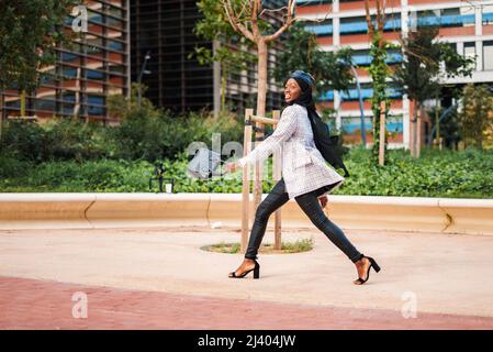 Side view full body of young stylish African American female in traditional headscarf swinging bag while strolling carelessly in urban park and looking away Stock Photo