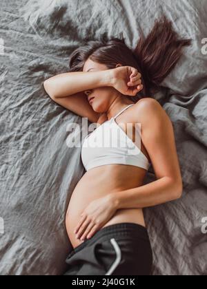 Tired pregnant woman sleeping on bed hiding face crying in depression or feeling morning sickness nausea. Mental health, fatigue, stress during Stock Photo