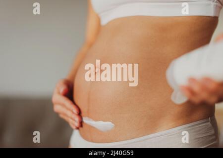 Pregnant woman putting cream on first trimester belly for stretch marks skin care prevention. Moisturizing dry skin during winter Stock Photo