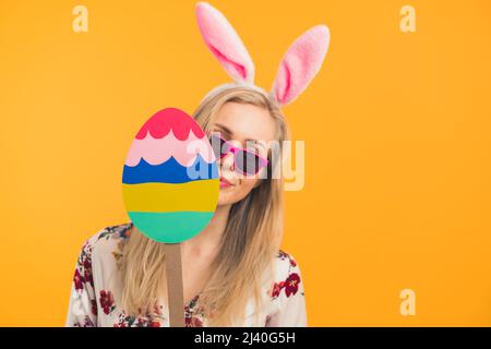 Happy Easter - a blonde young woman with Easter bunny ears holding a big wooden Easter egg holiday concept orange background medium closeup studio shot. High quality photo Stock Photo