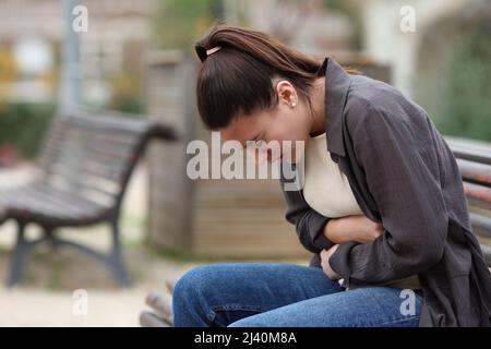 Profile of a teenage female suffering belly ache sitting on a bench in a park Stock Photo