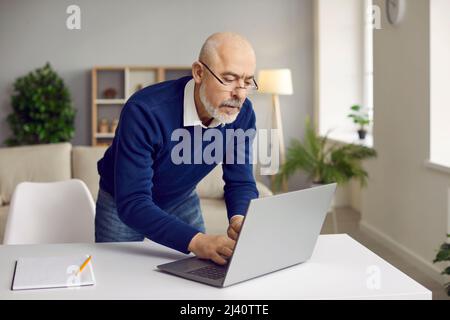 Serious senior businessman in glasses standing at his working desk and using his laptop Stock Photo