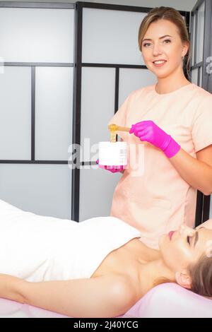 Sugaring. The beautician takes the sugar paste from the jar with a spatula. Lying woman customer waiting for sugaring procedure in the beauty salon Stock Photo