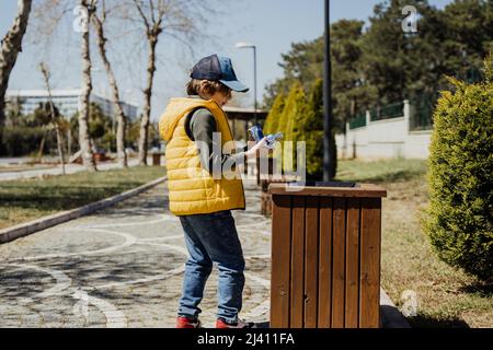 Schoolboy kid throwing the trash into dumpster. Boy using recycling bin to throw away the litter. Caucasian child recycles the junk into the trash-can Stock Photo