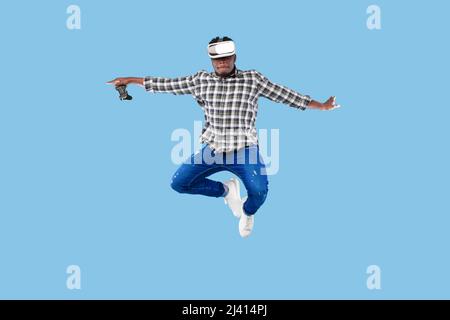 Young African American guy in VR headset holding joystick, jumping up in air on blue background Stock Photo