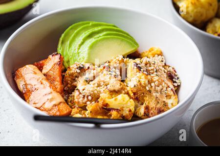 Vegan lunch bowl with baked cauliflower, sweet potato, avocado and peanut butter dressing, close-up. Healthy plant based food concept. Stock Photo