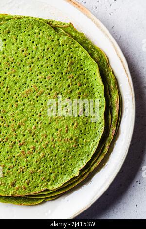 Stack of spinach crepes on white plate, top view. Green vegan pancakes, gray background. Healthy breakfast recipe. Stock Photo