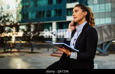 Successful businesswoman or entrepreneur taking notes and talking on cellphone outdoors. Stock Photo