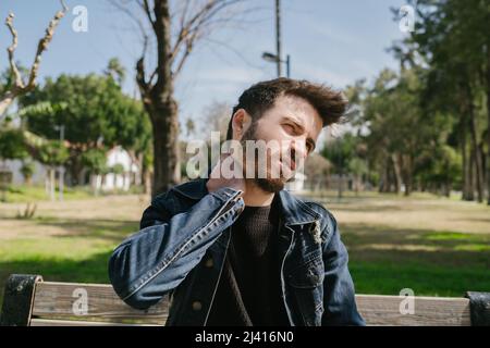 Young frustrated freelancer man is having a strong pain in neck. Sitting on a park bench at outdoor. Suffering, massaging it. Stock Photo