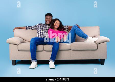 Happy millennial black couple relaxing on sofa, smiling at camera on blue studio background, full length Stock Photo