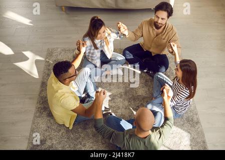Diverse group of friends sitting in circle on floor, holding hands and meditating together Stock Photo