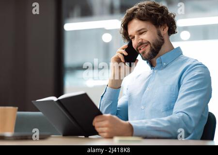 Smiling man talking on phone at office, reading diary Stock Photo