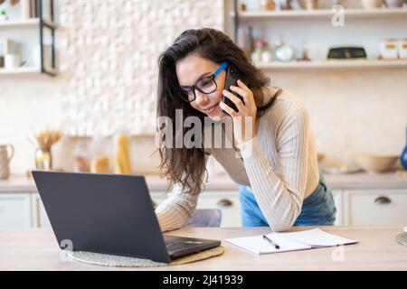 Freelance woman working from home, sitting at kitchen table, using laptop and having phone conversation Stock Photo